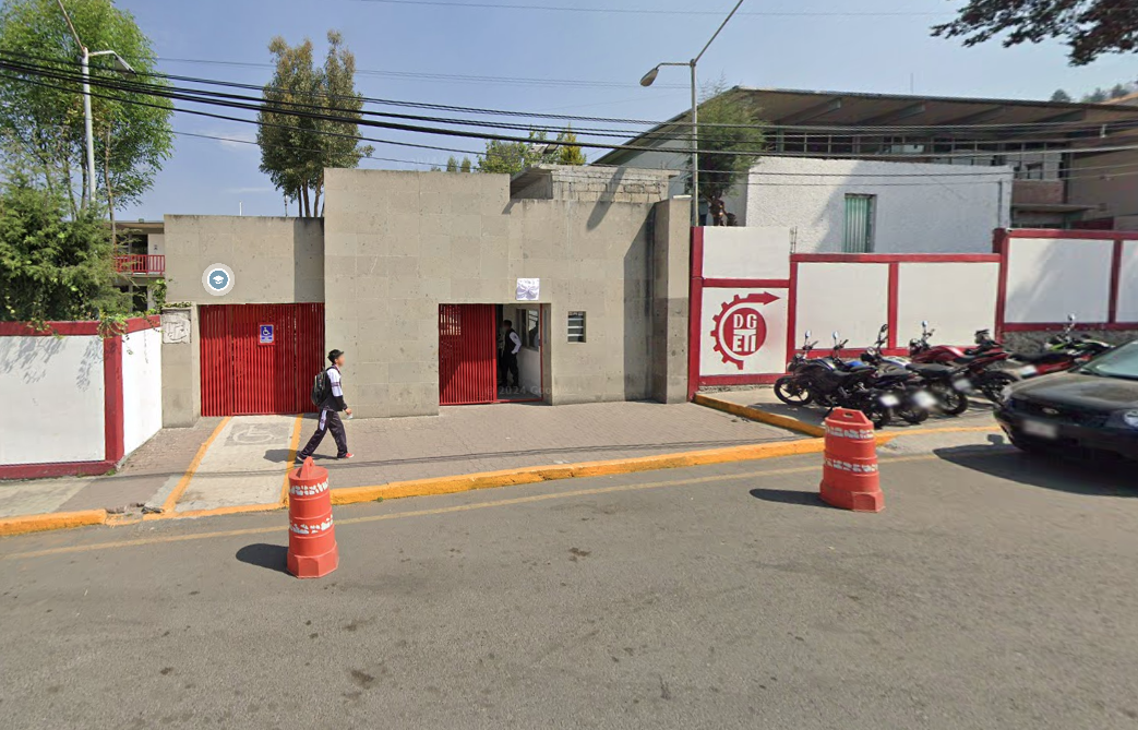 Fotografía en la que se muestra la entrada del plantel, conformada por muros de color gris. En medio de los muros hay dos portones de color rojo que dan acceso a las instalaciones, el portón del lado derecho tiene una rampa de acceso.